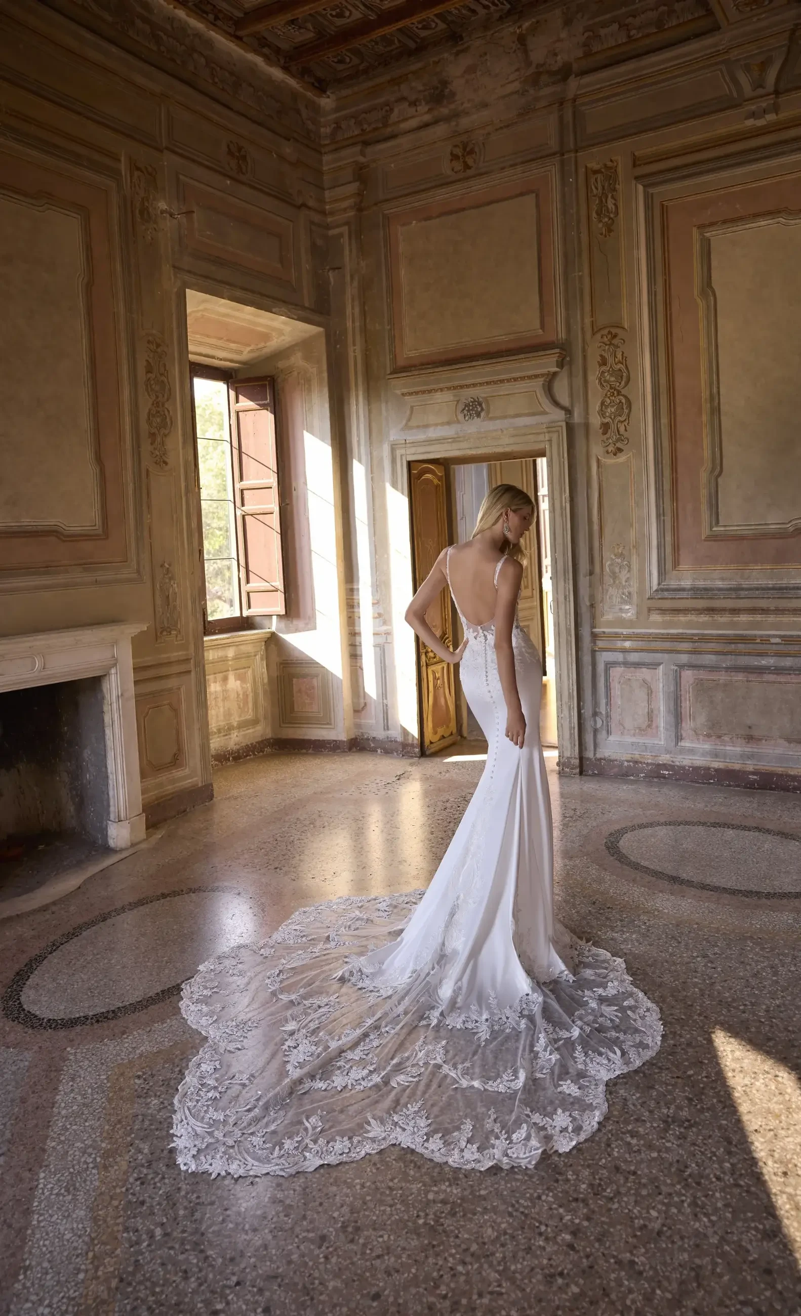 A woman in a sleeveless, lace-train wedding gown stands in a sunlit, ornate room with classic decorations, exuding elegance and serenity.