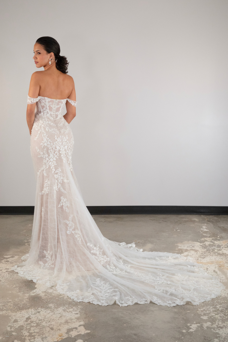 A woman stands facing away, wearing an off-shoulder lace wedding gown with a long train, in a minimalist setting. The moment feels elegant and serene.