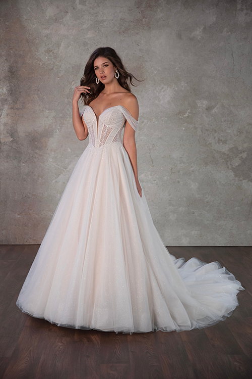 A woman stands in a light-filled room wearing an elegant, off-the-shoulder wedding gown with a full skirt and lace details, exuding grace and elegance.