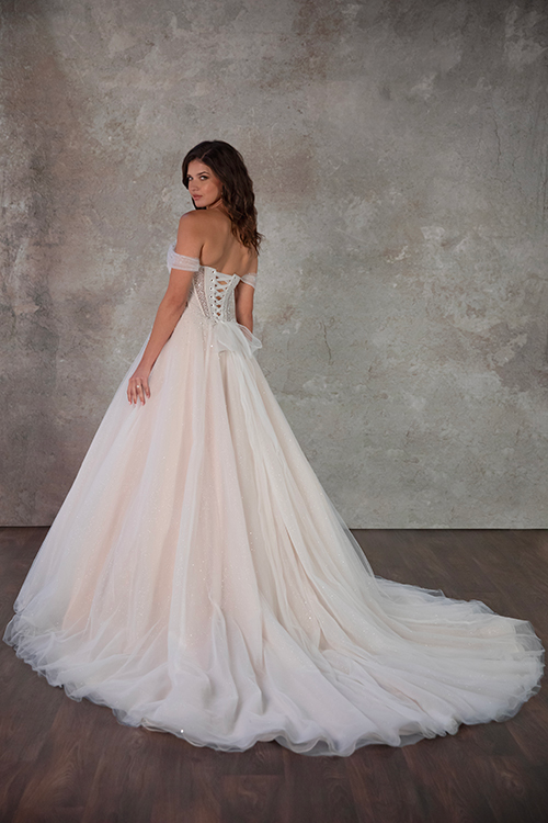 A woman in an elegant, off-the-shoulder white wedding gown stands with her back to the camera. The dress features a long, flowing train against a textured gray backdrop, conveying a romantic and graceful atmosphere.
