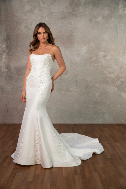 A woman wearing a fitted white strapless wedding gown with floral embellishments standing on a wooden floor against a textured gray background.