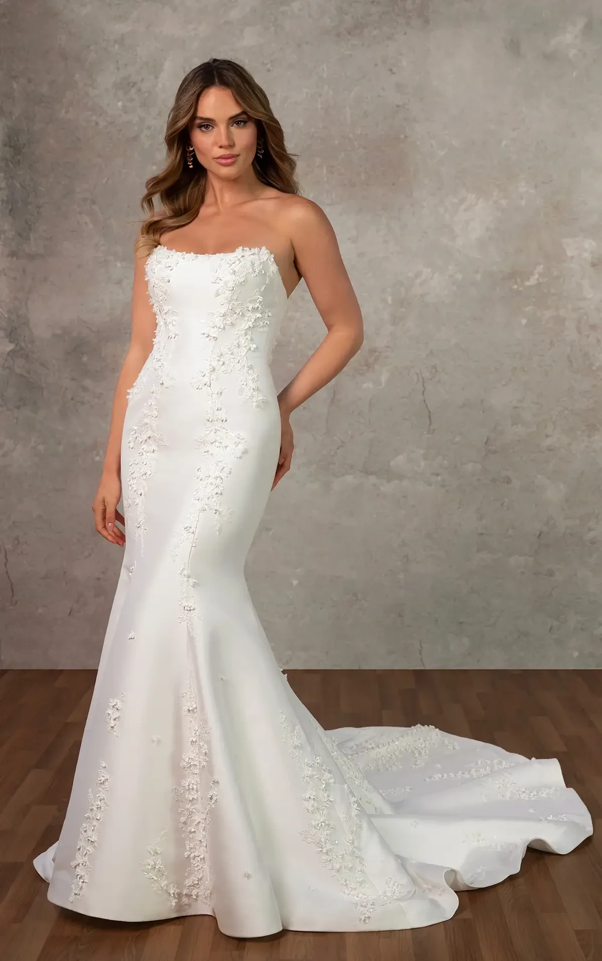 A woman in a fitted, strapless white wedding gown with floral embroidery stands on a wooden floor. She gazes confidently, exuding elegance against a textured gray backdrop.