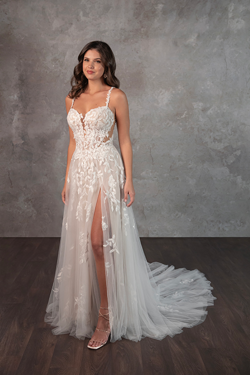 A woman in an elegant white wedding dress with lace details and a thigh-high slit stands on a wood floor, against a textured gray backdrop, exuding confidence.