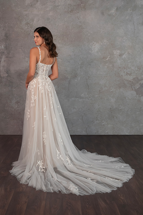 Bride in an elegant white gown with floral lace and a long train, standing on wooden floor. Her back is turned, exuding grace and serenity.