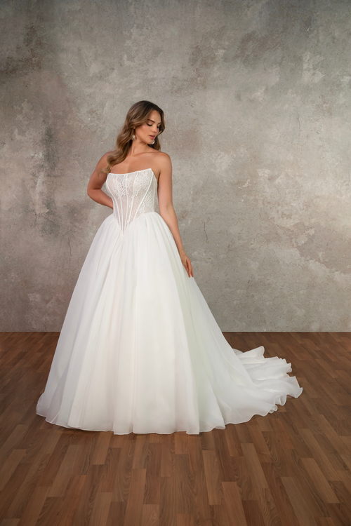 A woman in a strapless white wedding dress stands gracefully, showing off the full skirt and elegant design, with a textured gray background.