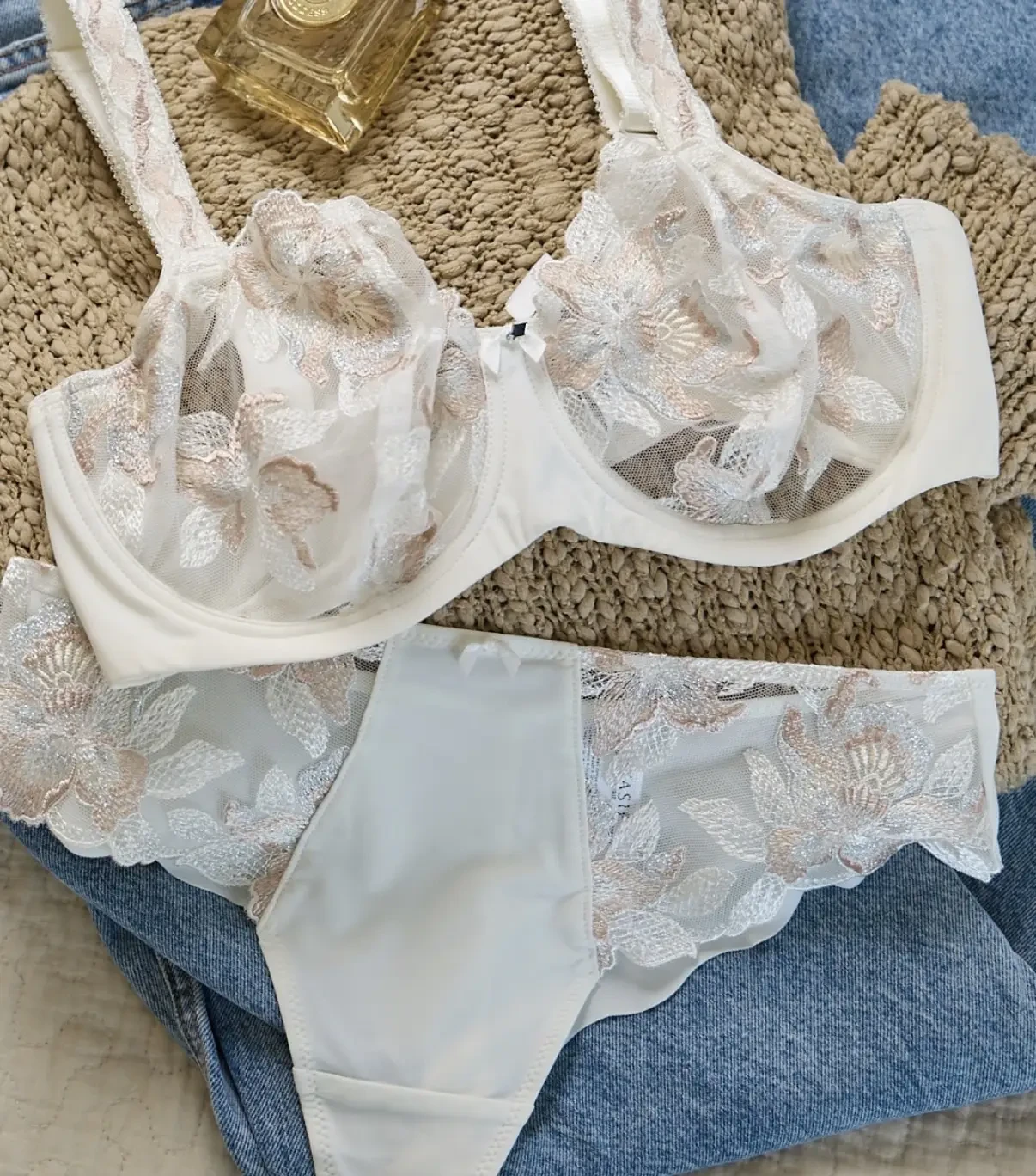 A cream-colored lingerie set featuring a lace bra with floral designs and matching panties, placed on a textured surface with a woven fabric and denim.