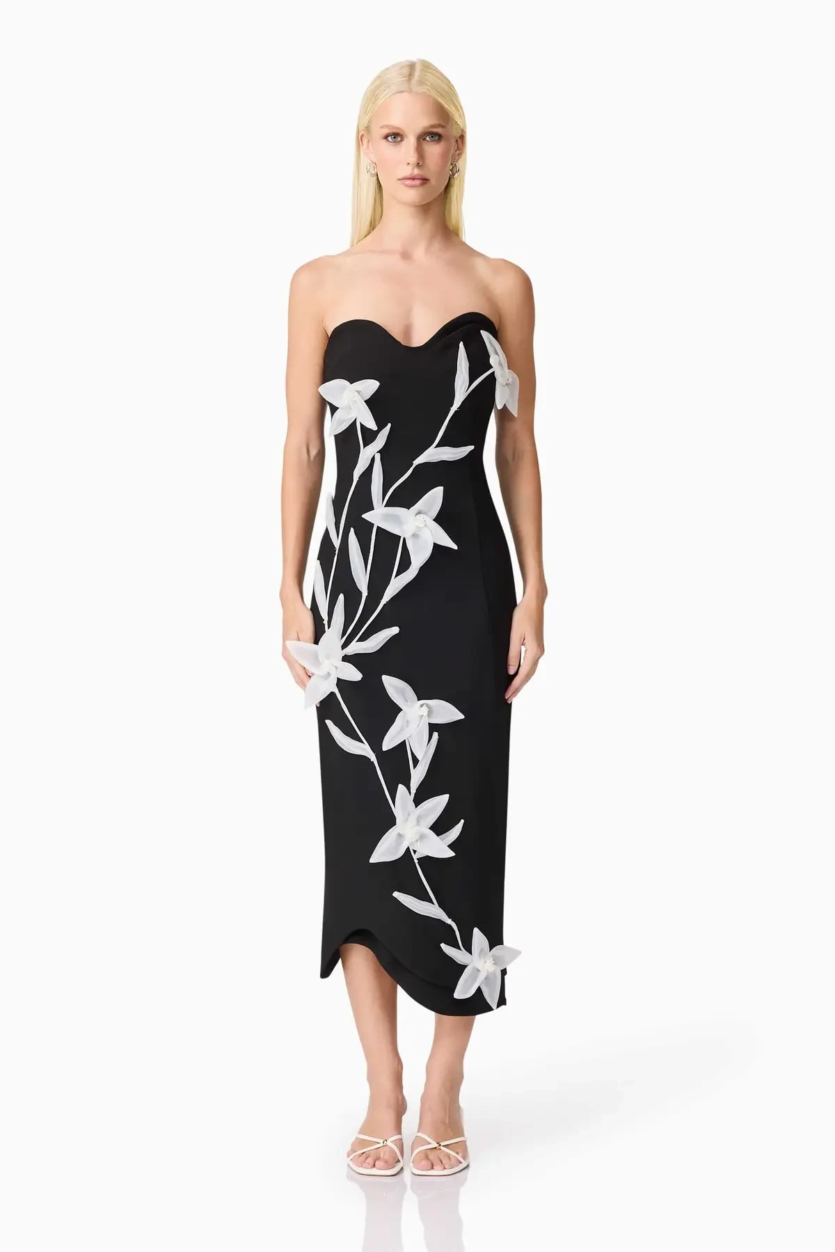 Model is wearing a romantic strapless midi dress with a sweetheart neckline, 3D floral detailing, and a sculptural silhouette that balances elegance and comfort.