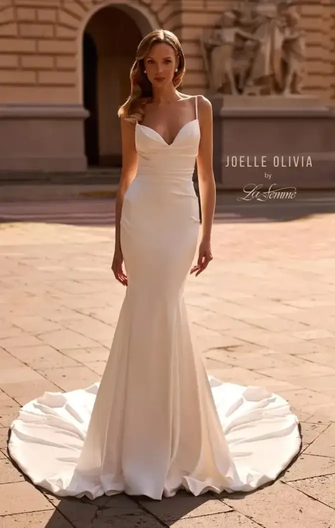 A model wearing a fitted white gown with a mermaid silhouette and a train, standing on a stone pavement in an outdoor setting.