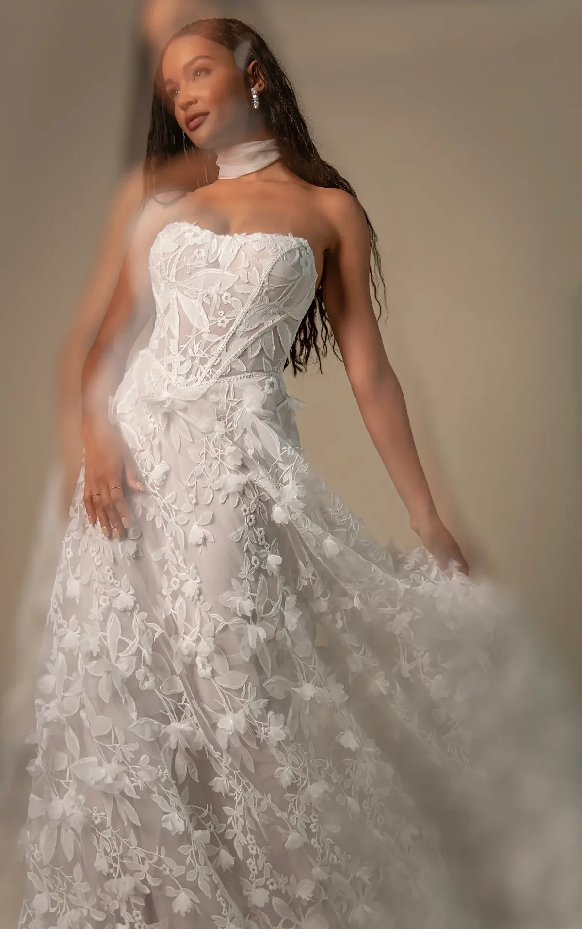 Model is wearing a strapless A-line gown with a corset bodice, soft scoop neckline, and a skirt adorned with 3D floral appliqués.