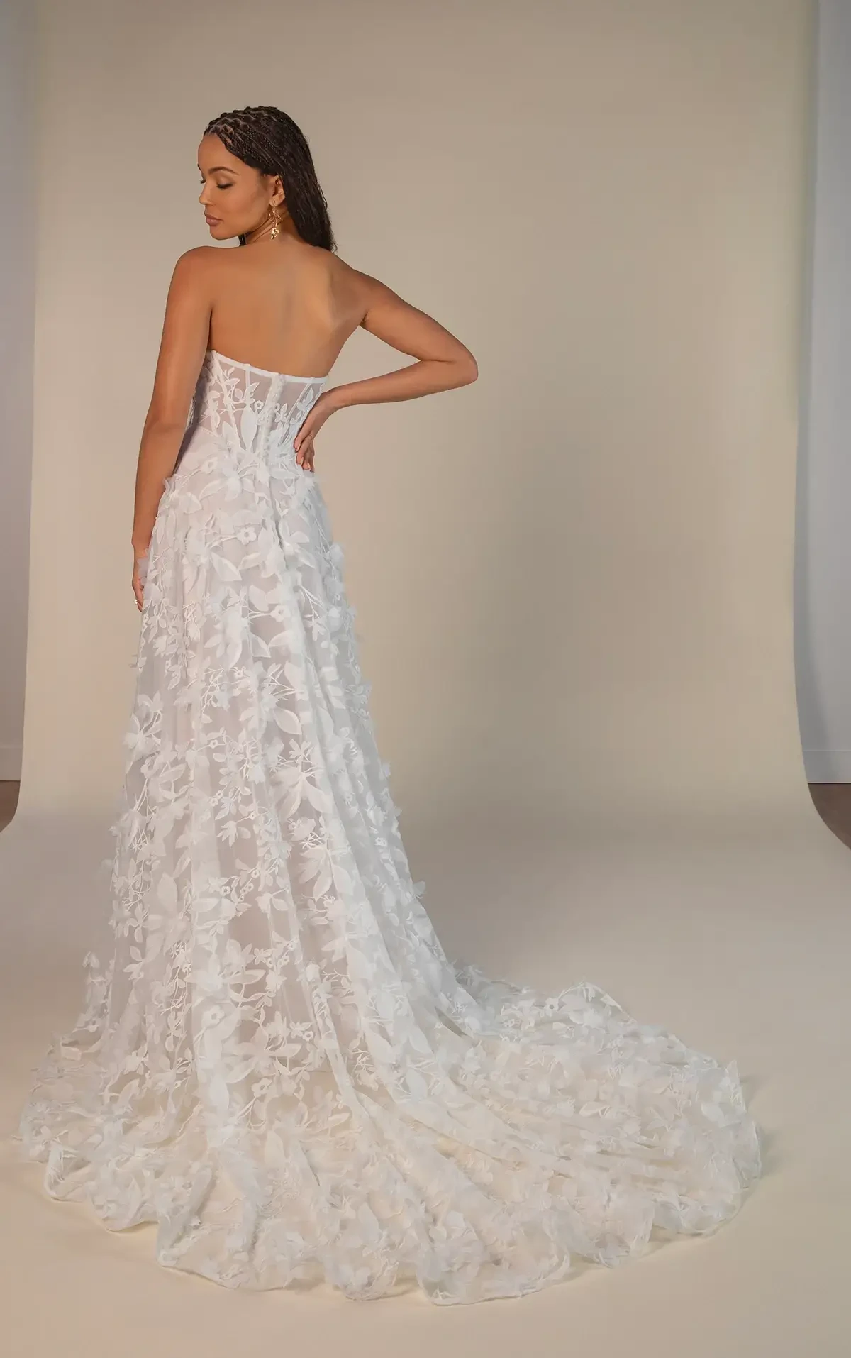 Model is wearing a strapless A-line gown with a corset bodice, soft scoop neckline, and a skirt adorned with 3D floral appliqués.