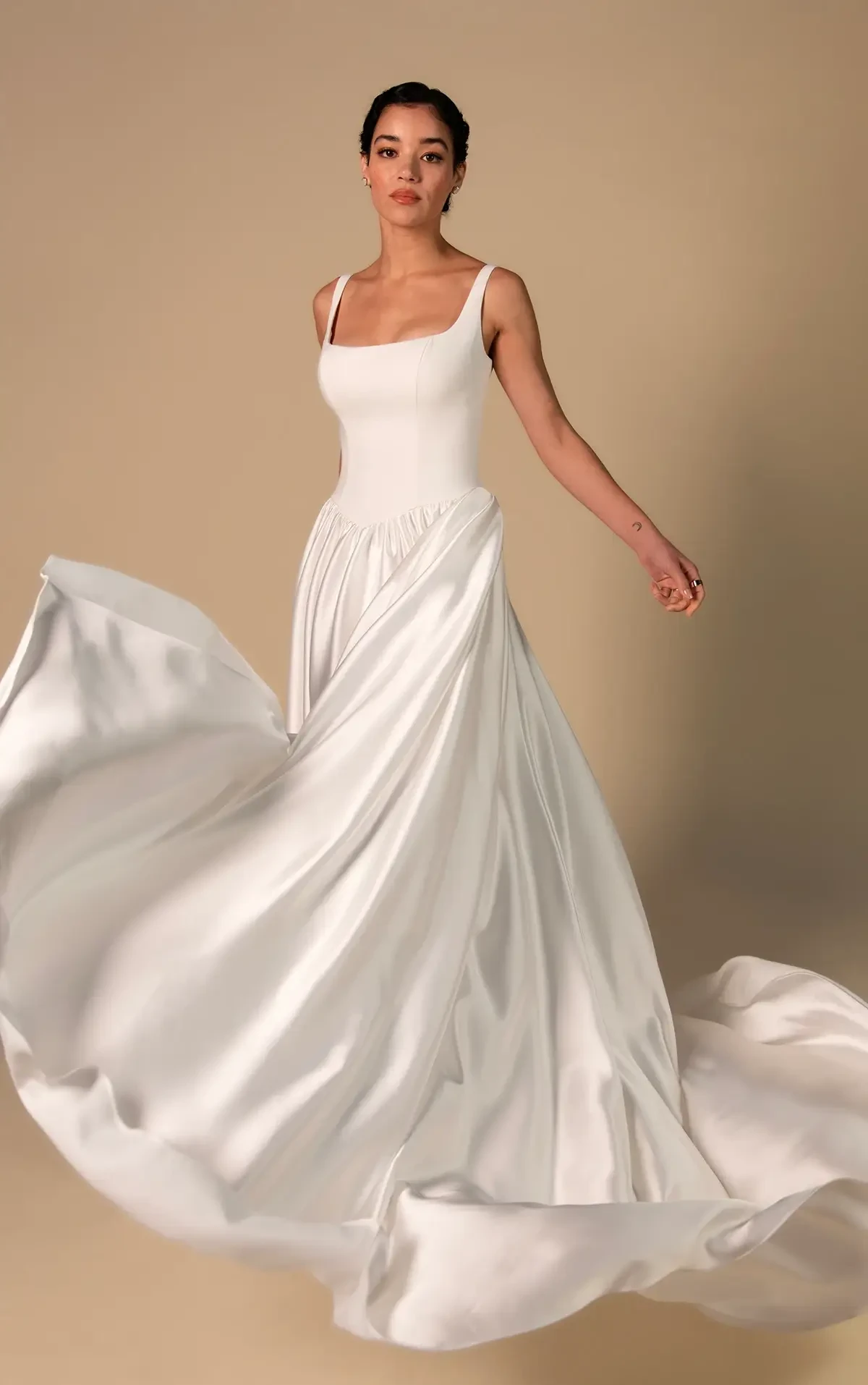 Model is wearing a sleek A-line gown with a square neckline, delicate straps, and a softly gathered skirt that flows gracefully.