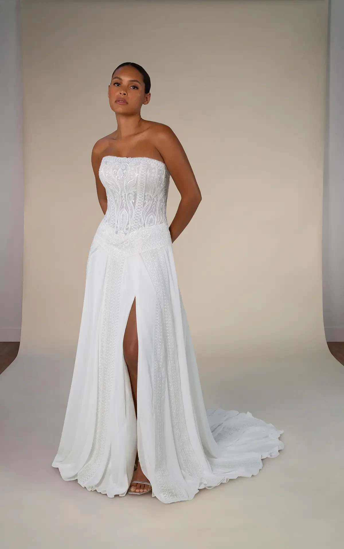 A woman in a strapless white wedding dress with lace detail and a front slit stands against a neutral backdrop, exuding elegance and confidence.