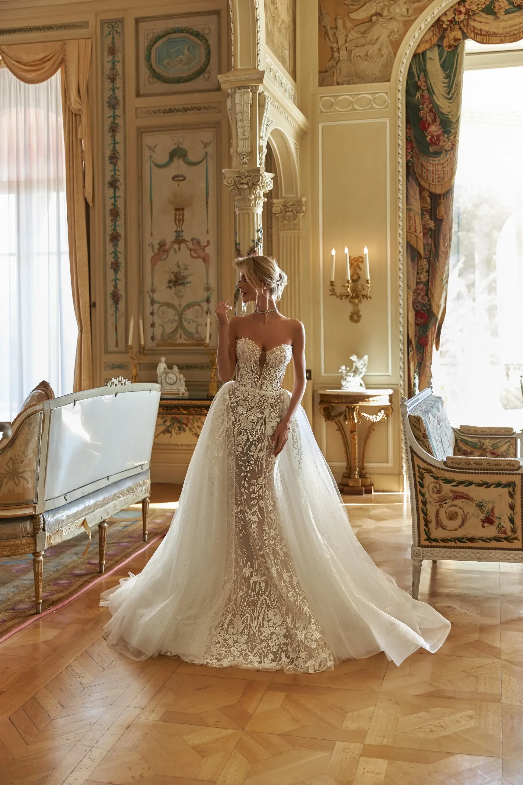 A woman in an ornate, elegant room wears a lace and tulle bridal gown, standing gracefully on a polished wooden floor with classic decor in the background.