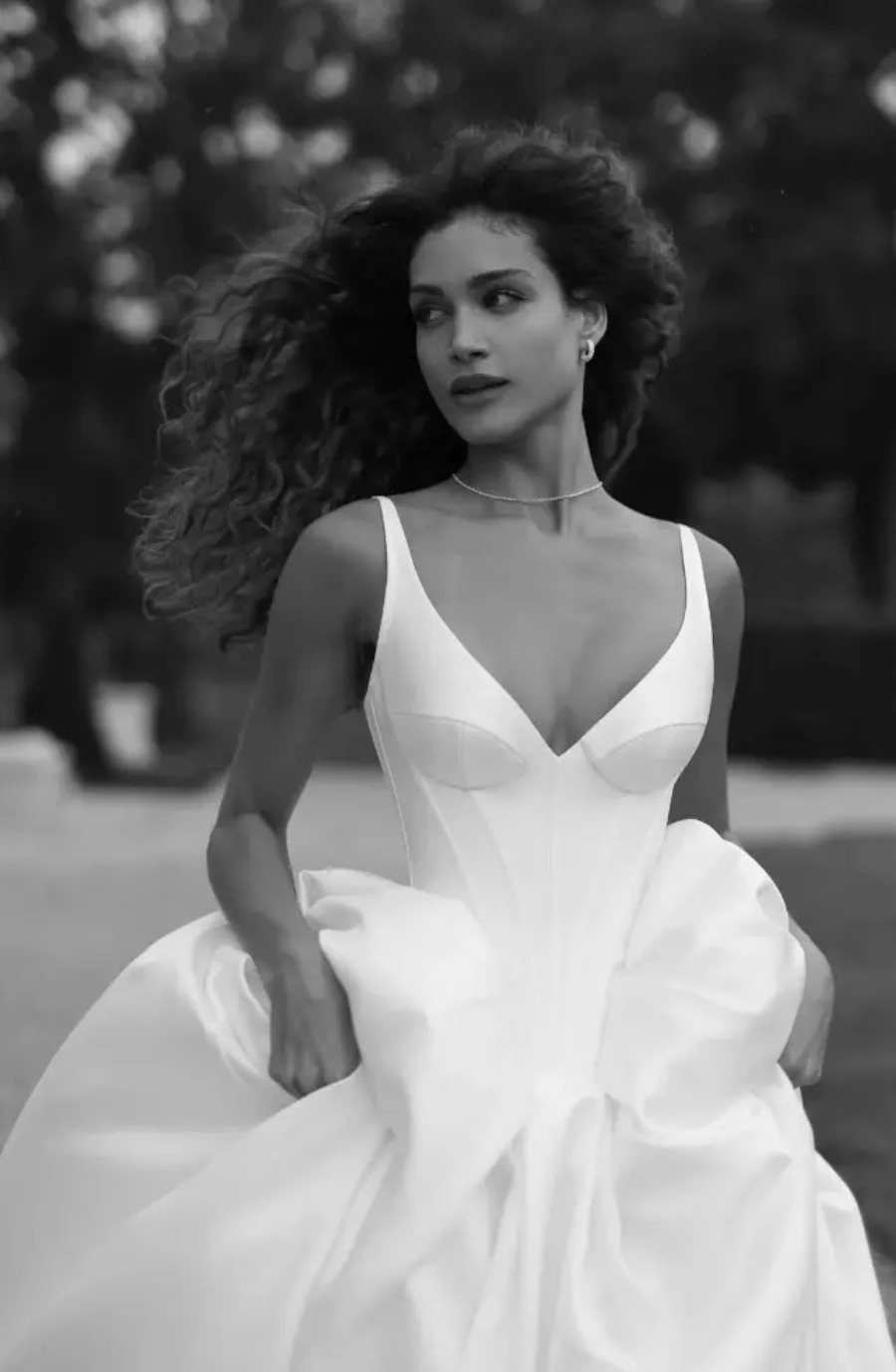 A woman in a flowing white dress with a deep V neckline, surrounded by nature in a black and white setting.