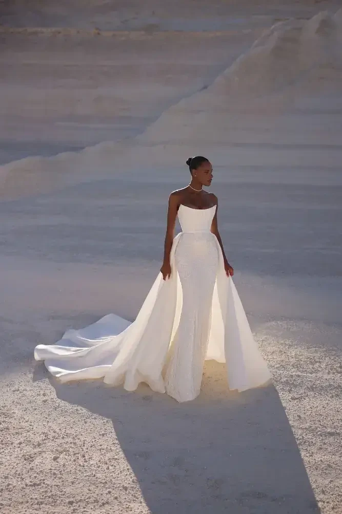 A woman wearing a stunning white gown stands in a vast, arid landscape. The dress features a fitted bodice and a flowing train, complementing her elegant pose.