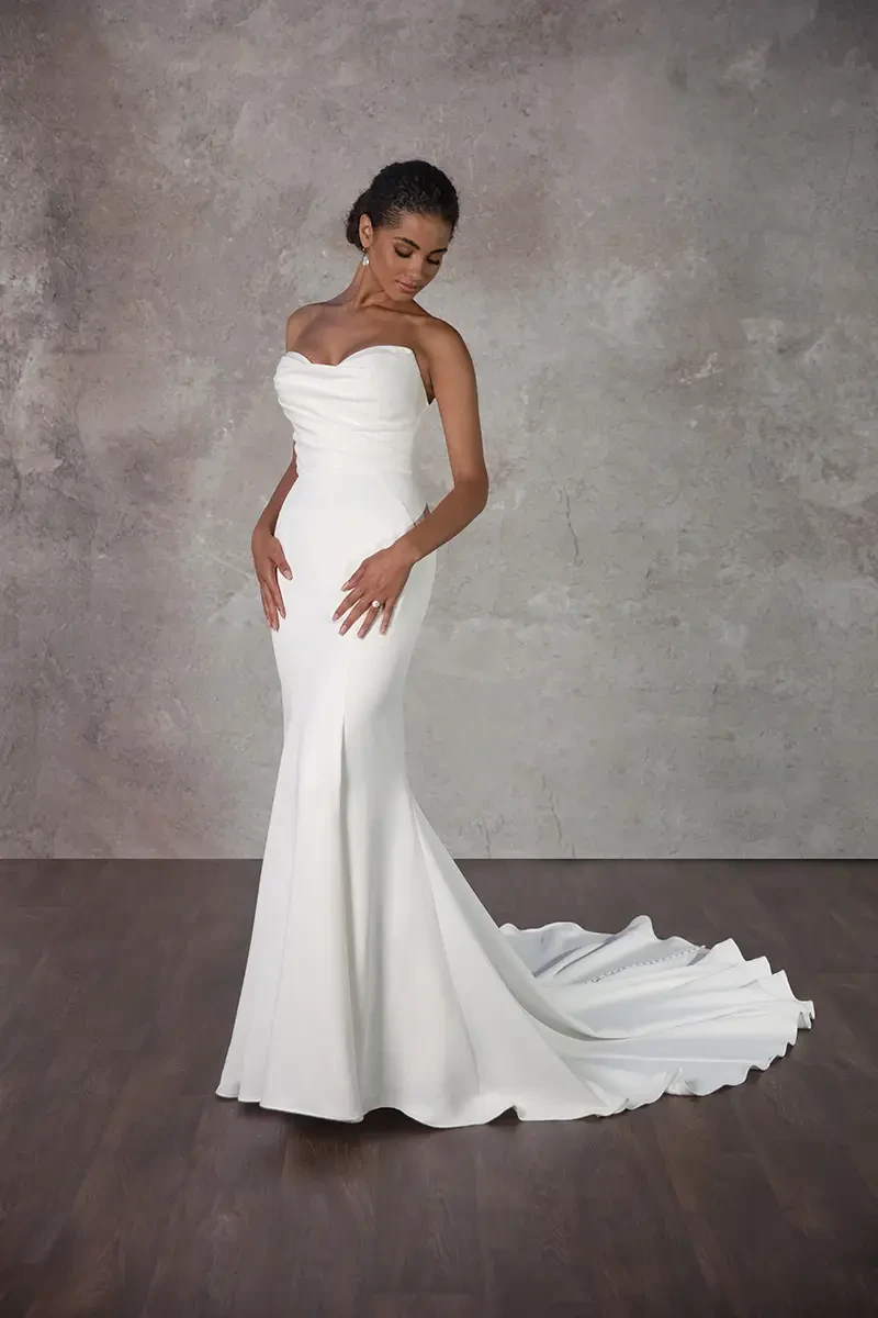 A woman in an elegant white strapless wedding gown poses gracefully. The dress features a fitted bodice and flowing train, against a textured gray backdrop.