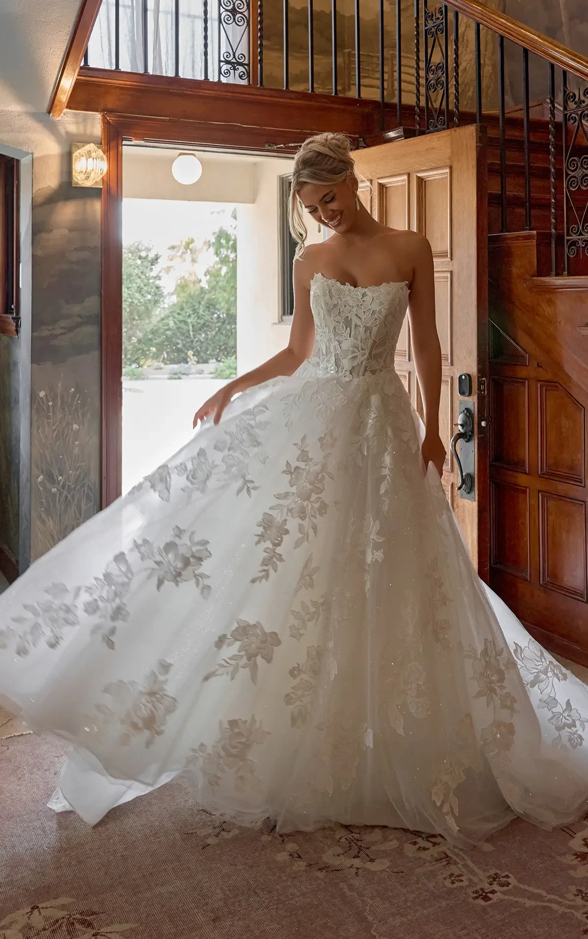 Bride wearing an elegant, floral lace wedding gown twirls gracefully in a warmly lit room. The scene feels joyful and serene, capturing a special moment.