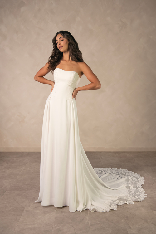 A woman wearing a strapless white wedding dress, posing confidently with her hands on her hips, against a neutral background.