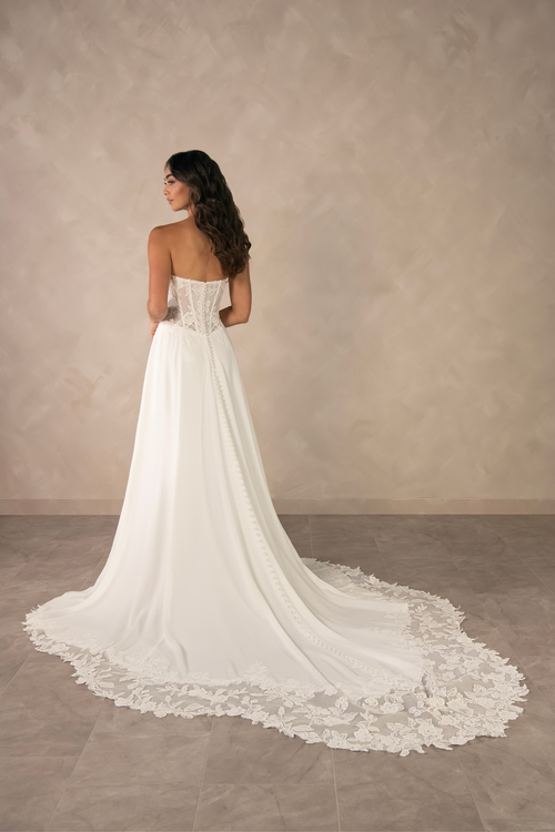 A woman is standing with her back to the camera, wearing a strapless wedding dress with a long train that has intricate lace detailing. The background is a neutral, textured wall.