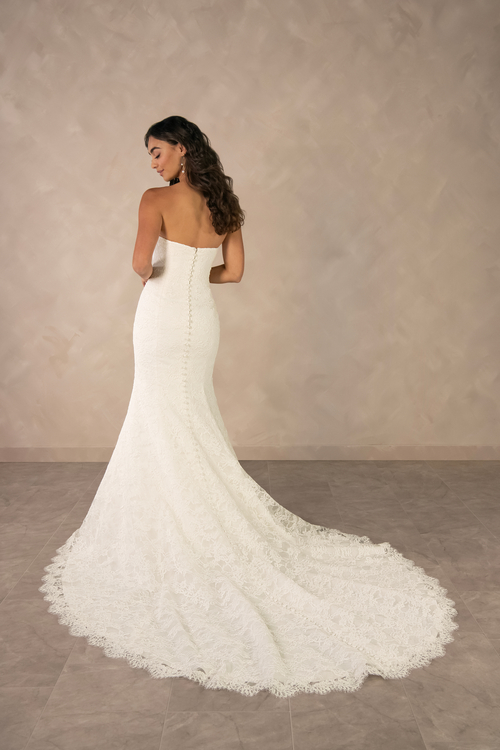A woman in a strapless white lace wedding dress, showing the back view with a long train, standing against a beige wall.
