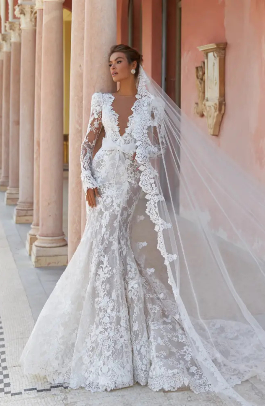 A bride poses in a stunning lace wedding gown with long sleeves and a dramatic train, complemented by a flowing veil, standing against a backdrop of columns and ornate architecture.