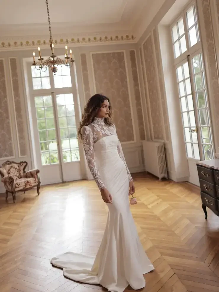 A woman in a white wedding dress with long lace sleeves stands in an elegant room with large windows and stylish decor.