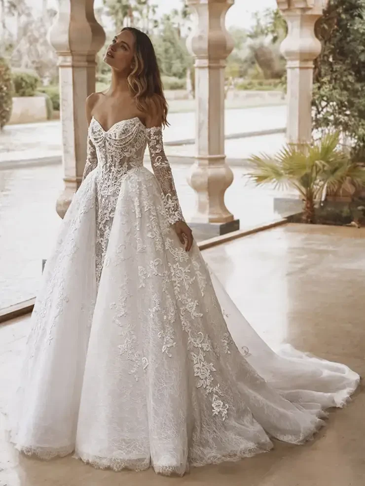 A bride in a stunning white wedding dress with intricate lace detailing, standing elegantly in a picturesque outdoor setting.