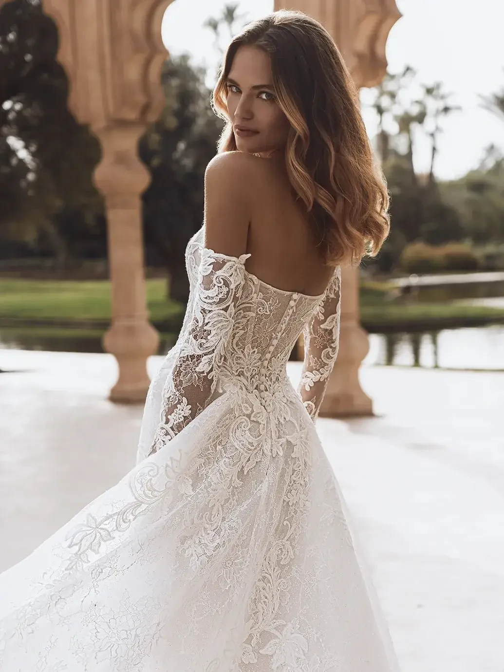 A woman in a detailed, off-shoulder lace wedding gown stands outdoors. She glances back with a serene expression, under an ornate stone arch.