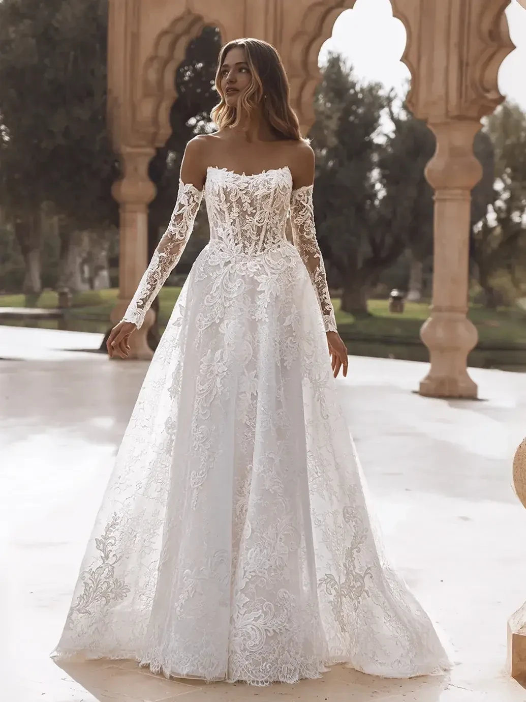 A woman in an elegant white lace wedding dress with long sleeves and a strapless neckline stands in a sunlit, arched stone courtyard, evoking a serene and romantic atmosphere.